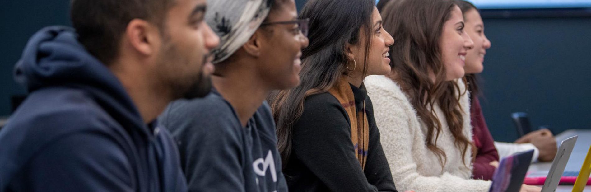 Undergraduate students attentively looking up from their laptops and facing the front of the classroom.